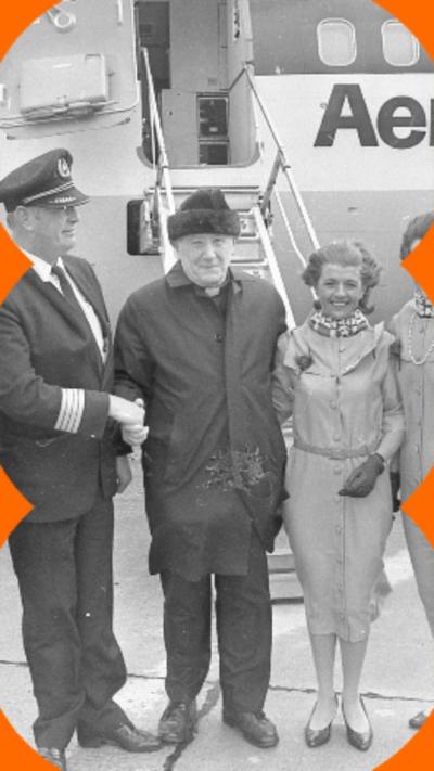 Black and white image of Monsignor James Horan stood stood in the middle of a group of pilots and air hostesses on an airport tarmac 