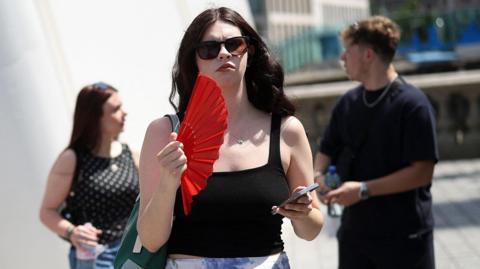 A woman on the street waves a red fan and holds her phone. She wears sunglasses and a black top.