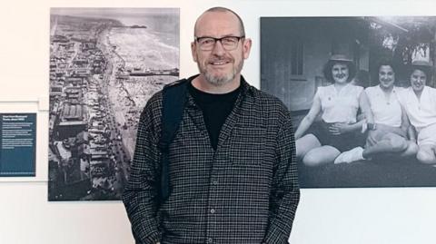 Steven Howells, who is wearing a shirt with a black T-shirt underneath and is smiling at the camera, is standing in front of a white wall with black and white photographs take by his grandad. The picture on the left is an aerial photograph of the coast and the image on the right is on three women sitting on the grass in front of a house. 