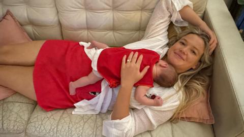 A woman with long blonde hair, wearing a white top and red skirt lying on a cream couch, with a baby sleeping on her chest. 