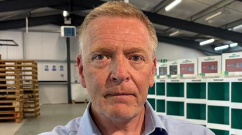 Andrew Moss sitting inside a warehouse area. He is looking straight down the camera and is wearing an open-necked blue shirt. He has light coloured hair and blue eyes. Behind him and on the right of the image are rows of empty white and green display cases. On the left of the image behind Andrew is a pile of empty wooden pallets. The warehouse has a pitched roof, held up by industrial metal girders, with strip lighting..