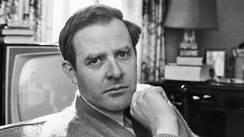A black and white portrait of John John le Carré sat in a living room, with a television behind him (Credit: Getty Images)