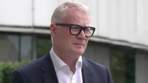 Richard Parker wears dark rimmed glassed and a dark suit jacket with a white shirt. He has short white hair.