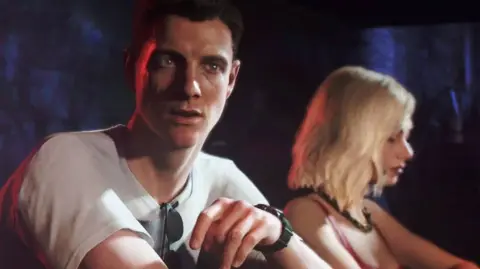 A screengrab from the new Bond video game. Two people are sitting close together in what looks like a warm, low‑lit bar or club. The lighting is mostly red and blue..  The person in the foreground (a young James Bond) is wearing a light shirt with sunglasses hanging from the collar, plus a wristwatch. They’re leaning slightly forward, relaxed, with their arm resting on the top of a bar.  Just behind them, a woman with light, shoulder‑length hair is wearing a sleeveless top and a necklace. She's turned slightly away from the camera, and the lighting catches their face more softly.