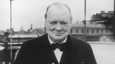 A black and white picture of Winston Churchill. He is looking directly into the camera. He is wearing a dark jacket, light shirt and a spotted bow tie. 