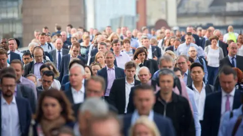 A large, dense crowd of commuters, mostly dressed in business and smart-casual attire, walking towards the camera on a city street or bridge, with buildings visible in the blurred background.