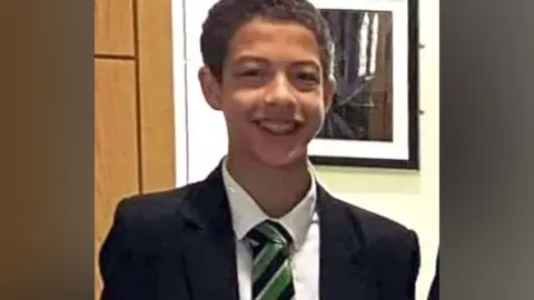 Noah Donohoe, a boy with dark hair, wearing a dark blazer, white shirt and green, black and white tie.