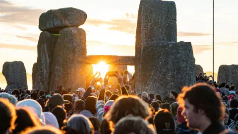 Heads in a crowd look towards the stones at Stonehenge. Some are holding up phone to take pictures as the sun peeks between the stones.