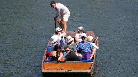 A group of people wearing sun hats sit in a wooden punting boat, floating on the water.