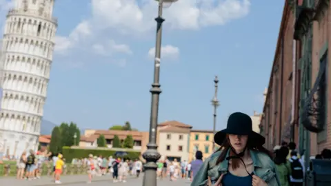 Getty Images A woman wearing a large sunhat and shielding herself from the sun walks past the leaning tower of Pisa
