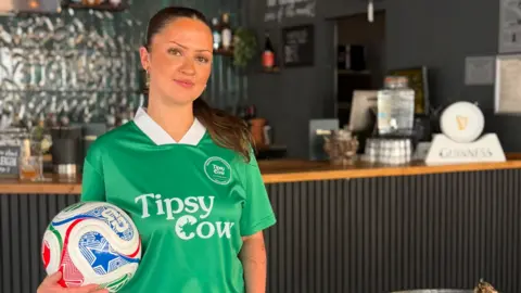 Freya, a woman with brown hair, wearing a green jersey holding a white and blue football, smiling for camera.