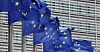 Blue European Union flags with yellow stars flutter in front of a modern glass office building.