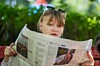 Woman holding and reading a newspaper outdoors with greenery in the background.