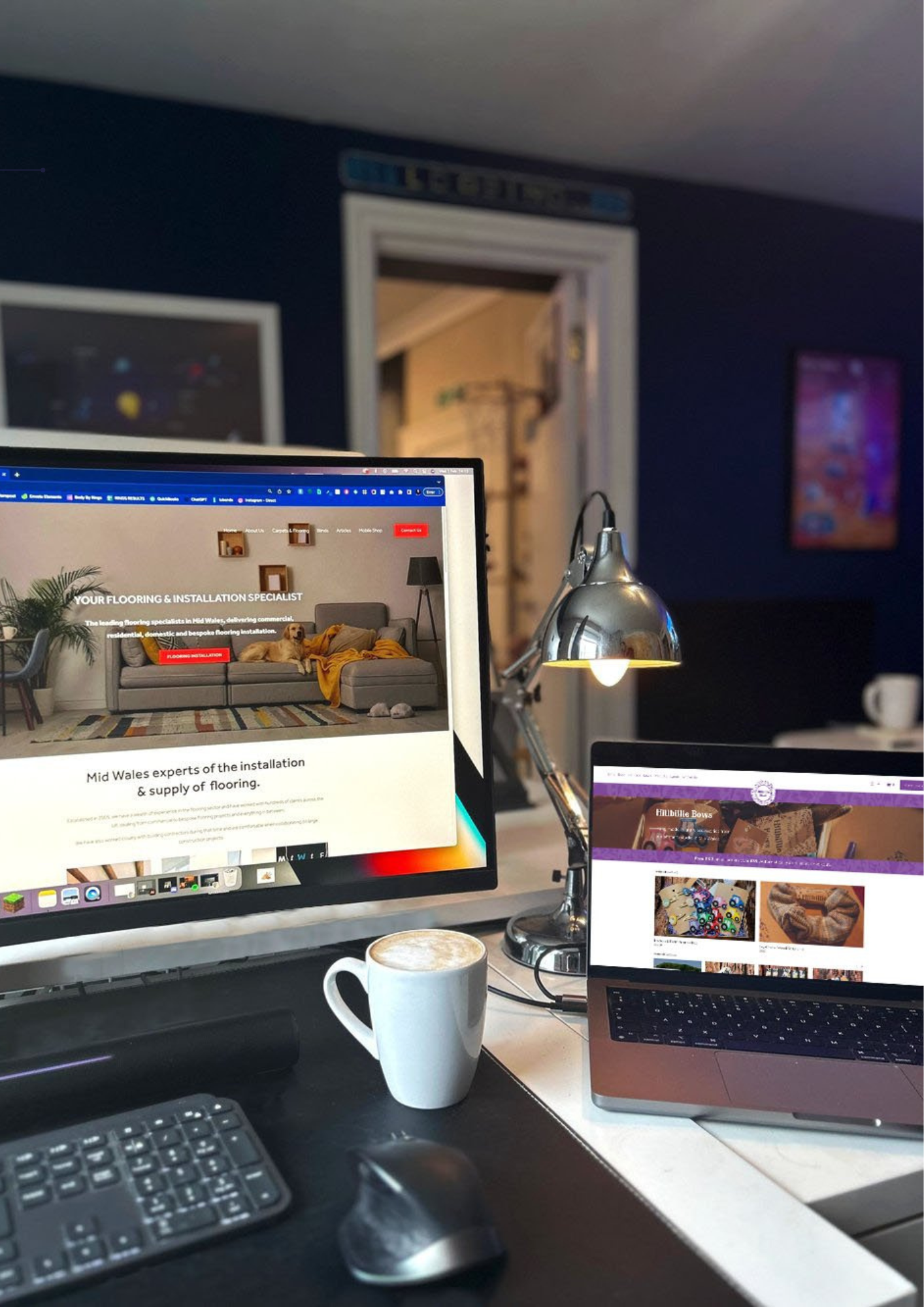 A home office desk setup with a monitor displaying a flooring company's website, a laptop showing a crafts website, a lamp, a white coffee mug with a latte, a keyboard, and a mouse. In the background, there is a door and a wall with a framed picture.