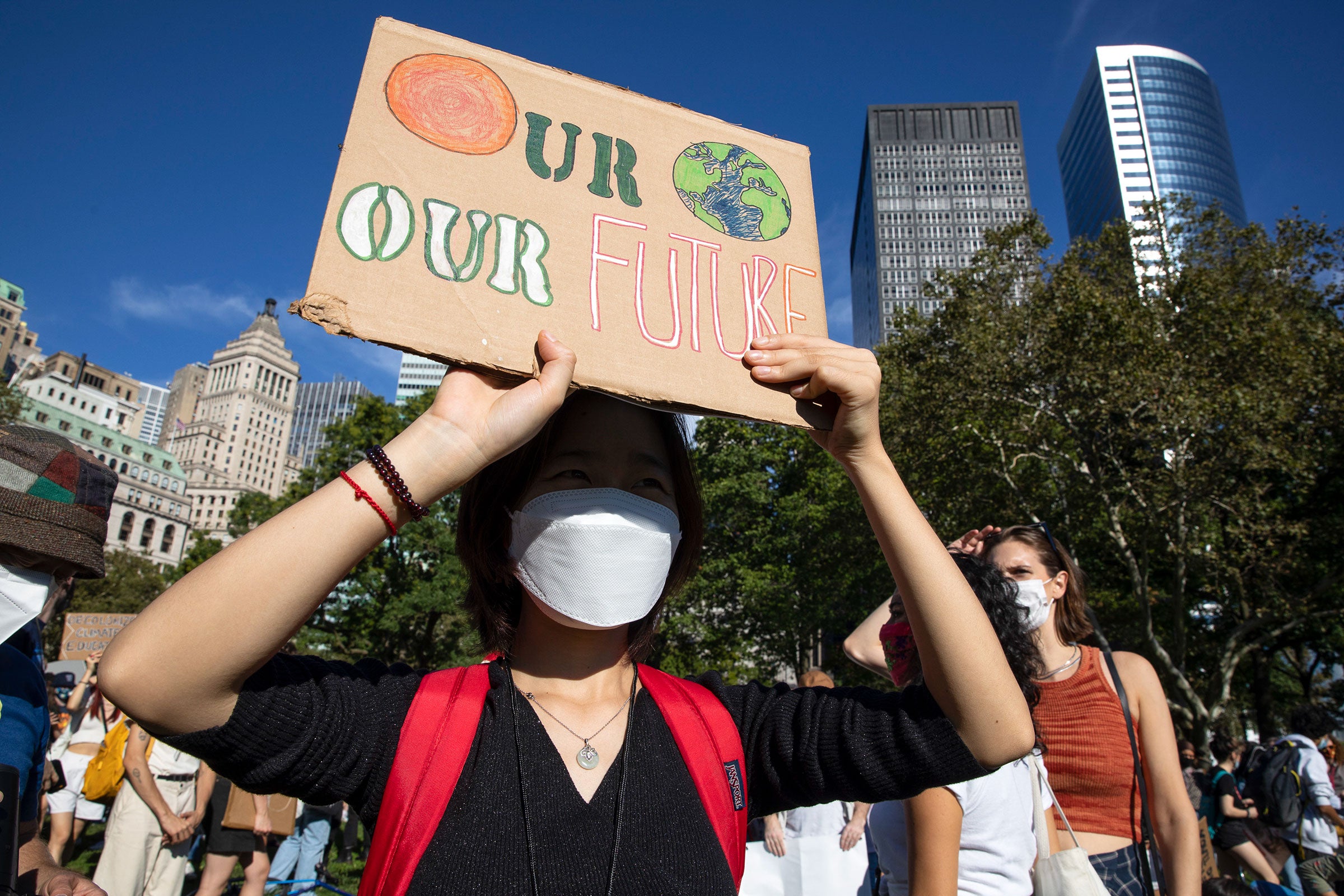 A woman wearing a face mask, black shirt, and a red backpack holds up a cardboard sign that says "Our Planet, Our Future" and the word "planet" is a painting of the Earth.