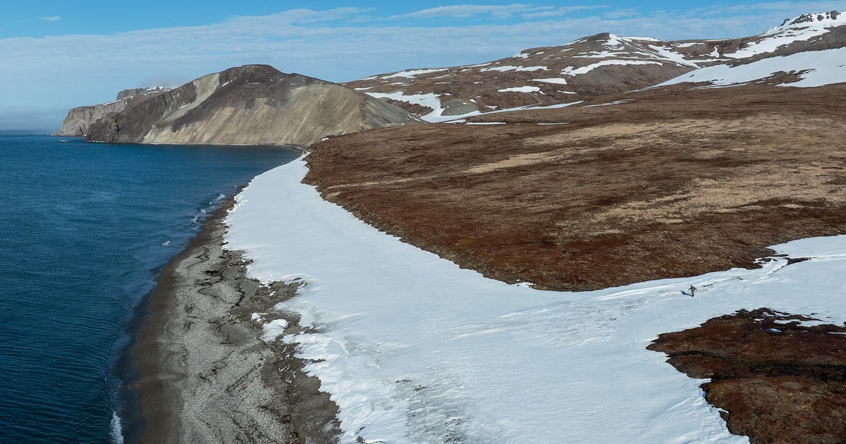 An aerial view of a snowy St. Matthew Island with a tiny spec of a human in the distance. 