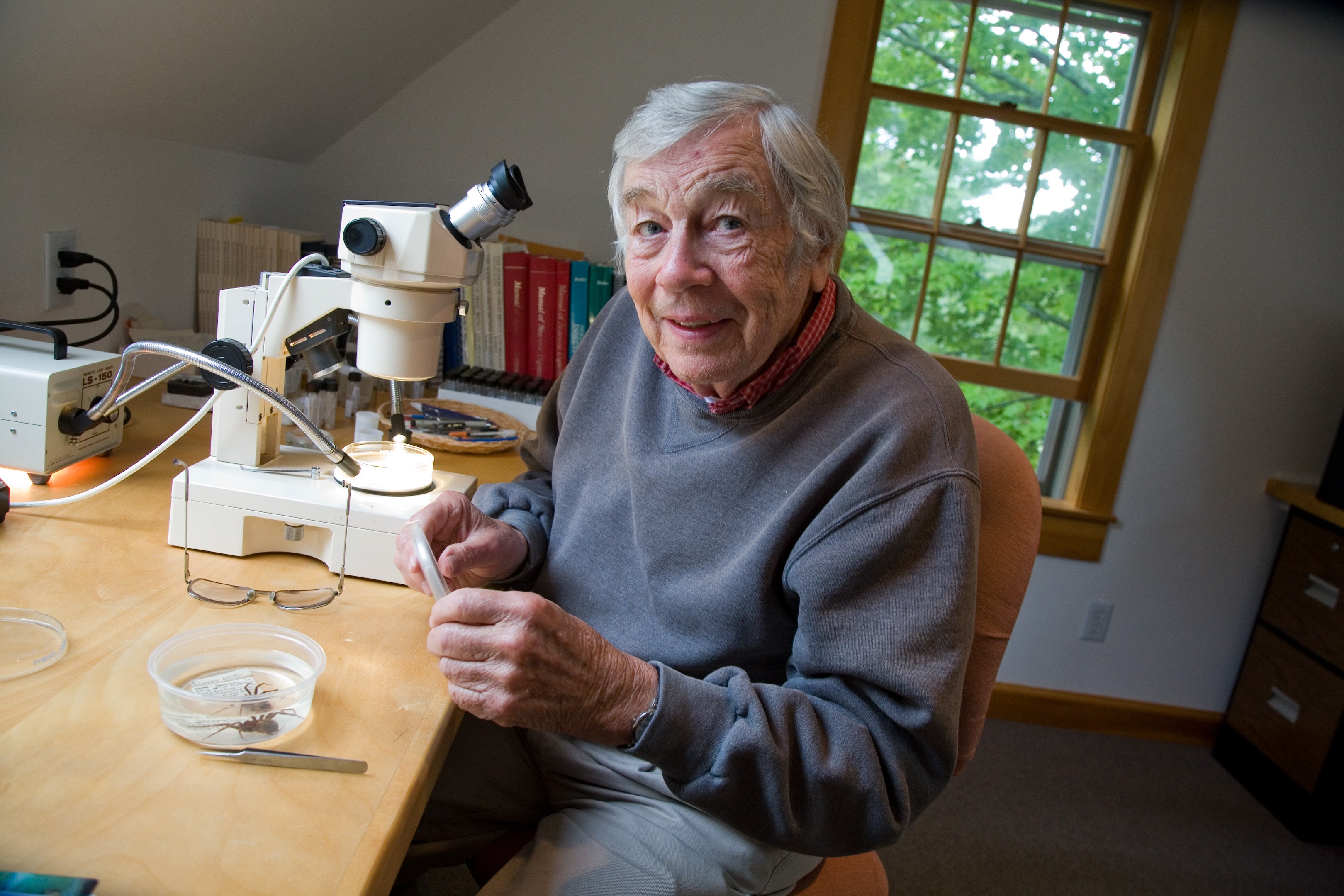 A man sitting at a desk with a microscope looks up and smiles.