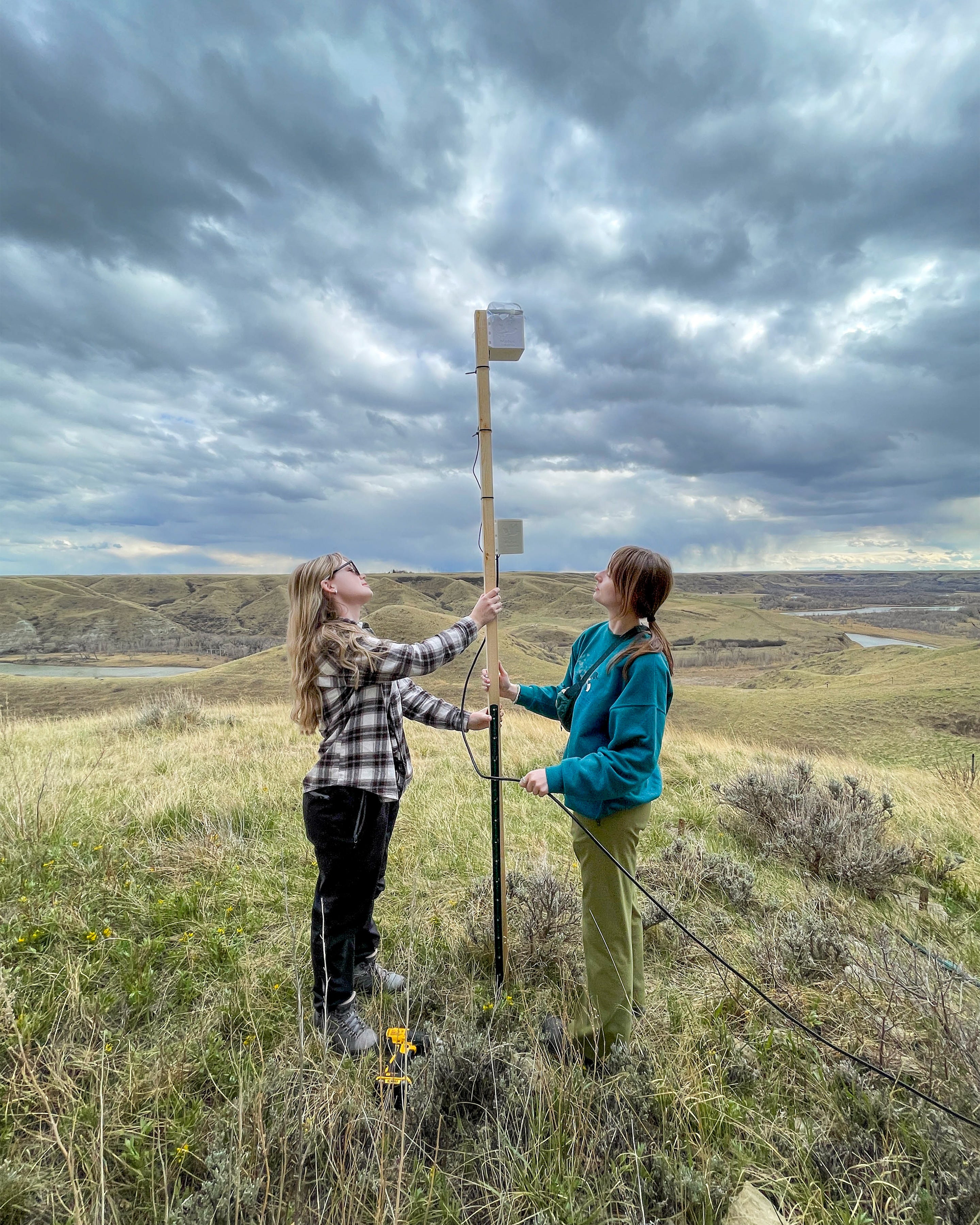 Two women installing boxes on a pole with an expansive green landscape behind them.