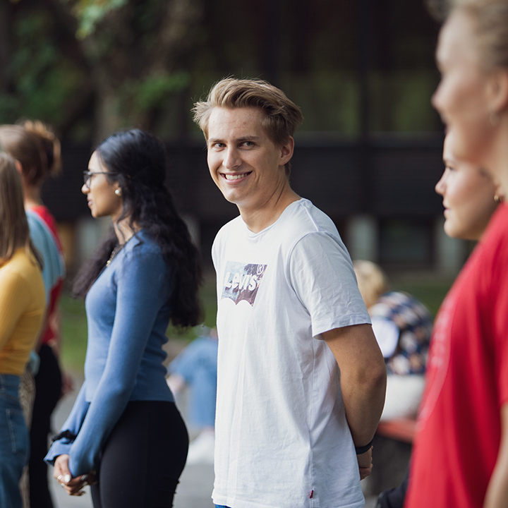 A male student smiles to the camera, with other students in the background.