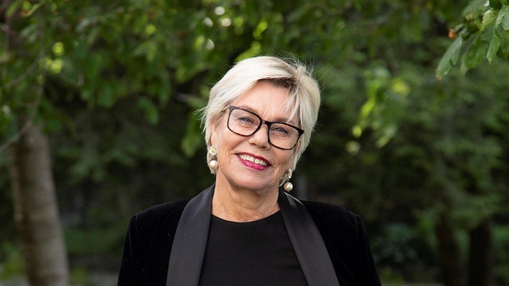 En eldre kvinne med lyst hår og briller ser rett frem. Smiler. 
