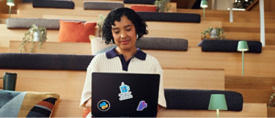 A curly-haired woman smiles while working on a sticker-covered laptop, seated on cozy steps with cushions and plants.