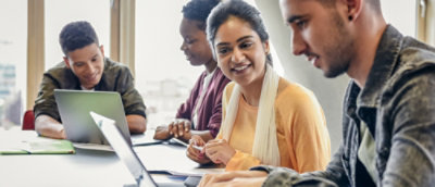 A diverse group of four students sits at a table with laptops, smiling and collaborating in a bright room.