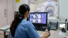 Pracowniczka ochrony zdrowia korzysta z komputera wyświetlającego skany mózgu. Widać pacjenta w aparacie MRI w tle, w szpitalnym otoczeniu.