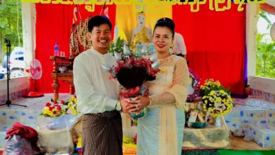 ကထိန်အလှူပွဲစာတန်းနောက်ခံနဲ့ ဘုရားဆင်းတုရှေ့မှာ ကထိန်သင်္ကန်းထည့်ထားတဲ့ ဖလားကို ကိုင်ပြီး ဓာတ်ပုံရိုက်ခံနေတဲ့ ဆရာမဒေါ်အေသီအောင်နဲ့ ခင်ပွန်း ဦးပြည့်ဖြိုးနိုင်