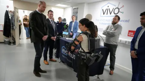 PA Media Prince William looks at a woman covered in an exoskeleton fitted with large battery packs. The prince wears a black suit and black sweater, A number of other people are looking on. Writing on the wall says VIVO. The UWE branding is  also visible. 